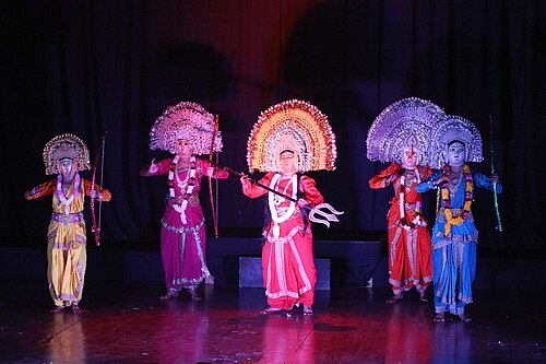 5 Chhau dancers with colorful costumes, large half-circle shaped hats, and weapons mid fight scene